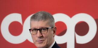 Portrait of a man in a dark suit and glasses, standing in front of a red backdrop with large white letters behind him.