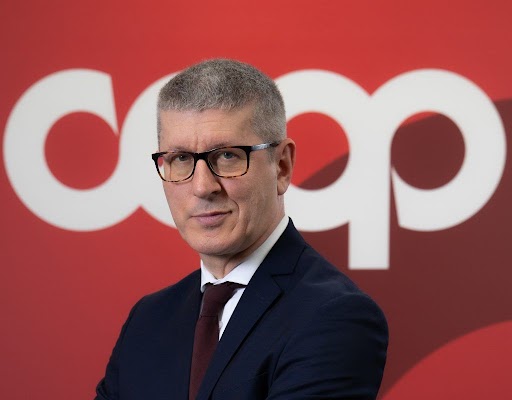 Portrait of a man in a dark suit and glasses, standing in front of a red backdrop with large white letters behind him.