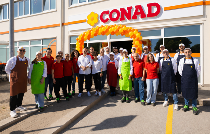 Group photo of Conad staff and volunteers under an orange balloon arch at a store opening, wearing colorful aprons and uniforms.