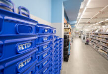 Blue stacked plastic bins labeled ACTION line the left foreground of a bright store aisle, with shelves of products visible on the right.