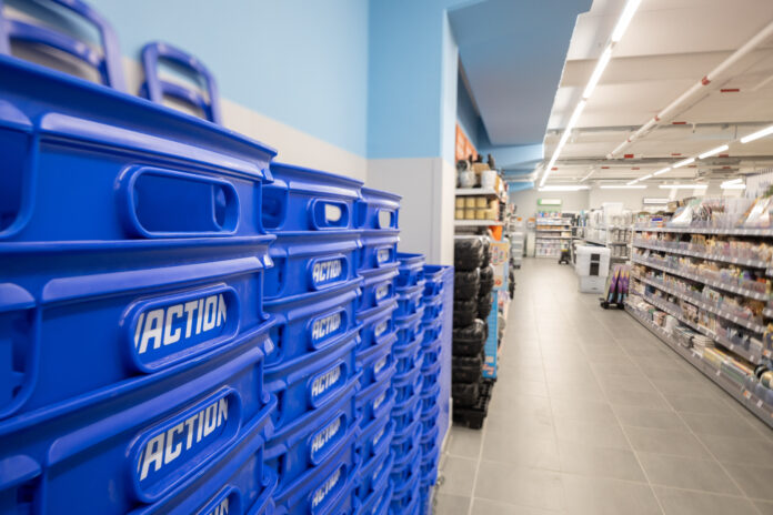 007_Action_repertorio_1 (1) Blue stacked plastic bins labeled ACTION line the left foreground of a bright store aisle, with shelves of products visible on the right.