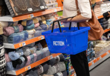 Action rafforza la presenza a Roma con il nuovo punto vendita Collatina Person with a brown bag pushing a blue shopping basket along a yarn aisle filled with colorful skeins and price tags on orange shelves in a store.