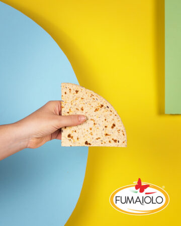 Hand holding a half tortilla chip against a bright blue circle and yellow background; FUMAIOLO logo in the bottom-right corner.