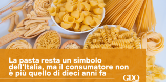 Assorted dry pasta shapes (penne, fusilli, farfalle, spaghetti) spread on a light surface with an orange banner containing Italian text about pasta as a symbol of Italy.