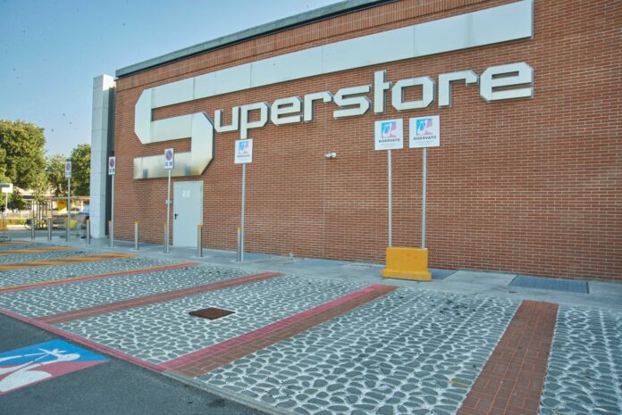 260728_071052 @ esselunga Exterior view of a brick building with a large white 'SUPERSTORE' sign on the wall, with handicap parking signs and bollards in the foreground.