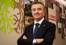 Conad Nord Ovest entra nel mercato drugstore con l’acquisizione di PiùMe Man in a dark suit and blue polka-dot tie stands with arms crossed in a modern lobby, a bright lime-green mural on the wall beside him.