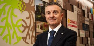 Conad Nord Ovest entra nel mercato drugstore con l’acquisizione di PiùMe Man in a dark suit and blue polka-dot tie stands with arms crossed in a modern lobby, a bright lime-green mural on the wall beside him.