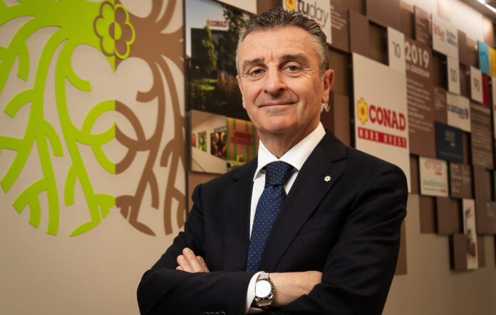 A.Adamo_Ascari 49.copia Man in a dark suit and blue polka-dot tie stands with arms crossed in a modern lobby, a bright lime-green mural on the wall beside him.
