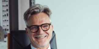 Smiling mature man with gray hair and glasses, wearing a dark suit and tie in an office setting.