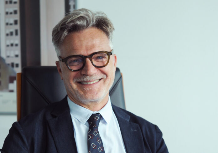 Alessandro Camattari3_Vert_Stamp Smiling mature man with gray hair and glasses, wearing a dark suit and tie in an office setting.