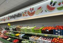 Supermarket produce aisle with colorful fruit illustrations on a banner above and fresh fruit/veg displays below.