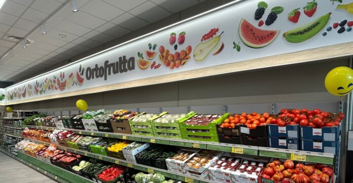 Apertura Fabrica Roma Supermarket produce aisle with colorful fruit illustrations on a banner above and fresh fruit/veg displays below.