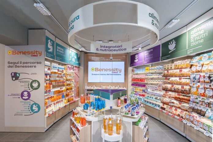 Benessity_4 Wide view of a bright health store aisle with Benessity branding, a central display table, and well-stocked shelves along both sides.