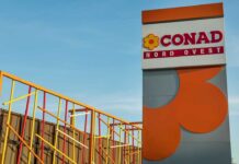 Conad Nord Ovest integra i primi negozi ex Realco nella rete di vendita Sign reads 'CONAD NORD OVEST' with a yellow flower logo, set beside a tall gray-and-orange architectural structure against a blue sky, and colorful metal railing artwork on the left—likely the exterior of a CONAD store site.