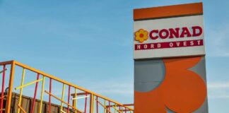 Conad Nord Ovest integra i primi negozi ex Realco nella rete di vendita Sign reads 'CONAD NORD OVEST' with a yellow flower logo, set beside a tall gray-and-orange architectural structure against a blue sky, and colorful metal railing artwork on the left—likely the exterior of a CONAD store site.