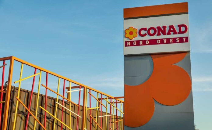 Sign reads 'CONAD NORD OVEST' with a yellow flower logo, set beside a tall gray-and-orange architectural structure against a blue sky, and colorful metal railing artwork on the left—likely the exterior of a CONAD store site.