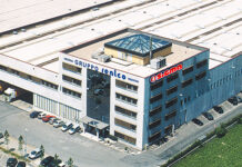 Aerial view of a multi-story office building with a blue facade and ground parking, surrounded by greenery and a large white roofed complex nearby.