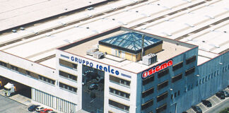 Aerial view of a multi-story office building with a blue facade and ground parking, surrounded by greenery and a large white roofed complex nearby.