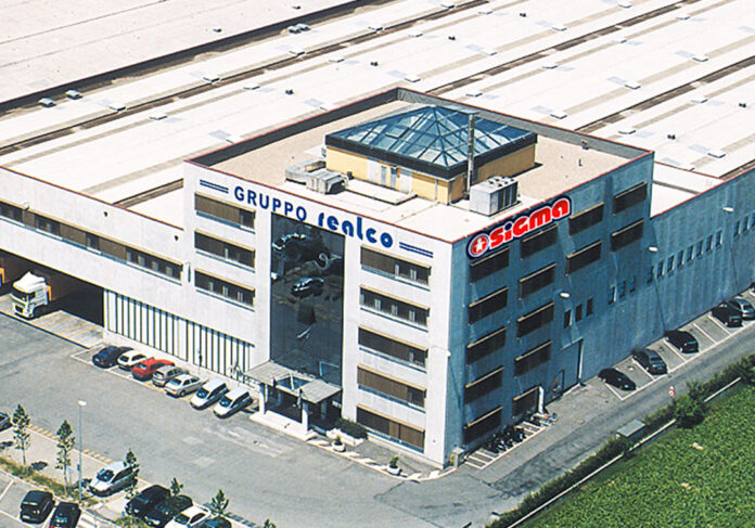 ChiSiamo_Header_Mobile2 Aerial view of a multi-story office building with a blue facade and ground parking, surrounded by greenery and a large white roofed complex nearby.
