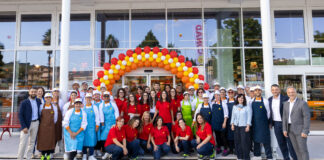 Group of staff and managers posing in front of a store entrance under a colorful balloon arch (red, yellow, orange) for a celebration event.