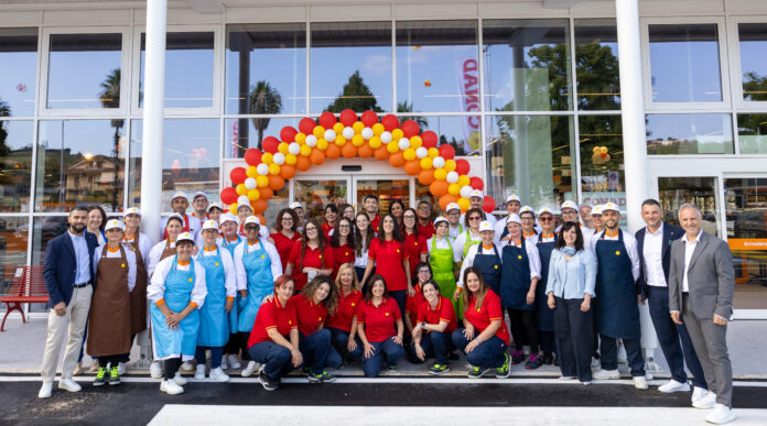 Colli del Tronto_3 Group of staff and managers posing in front of a store entrance under a colorful balloon arch (red, yellow, orange) for a celebration event.