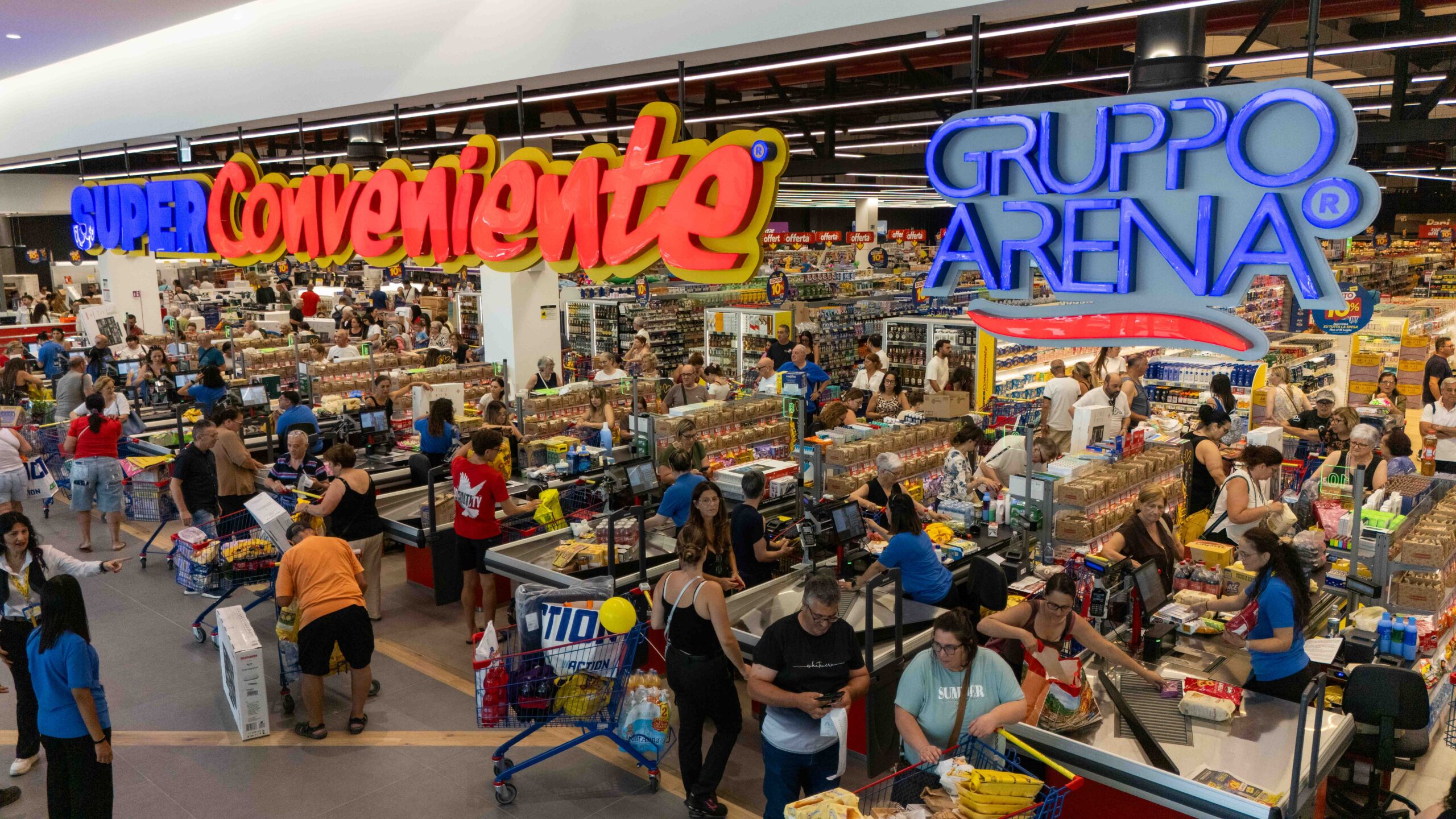 Busy supermarket checkout area with many customers at registers and a large, colorful overhead sign.