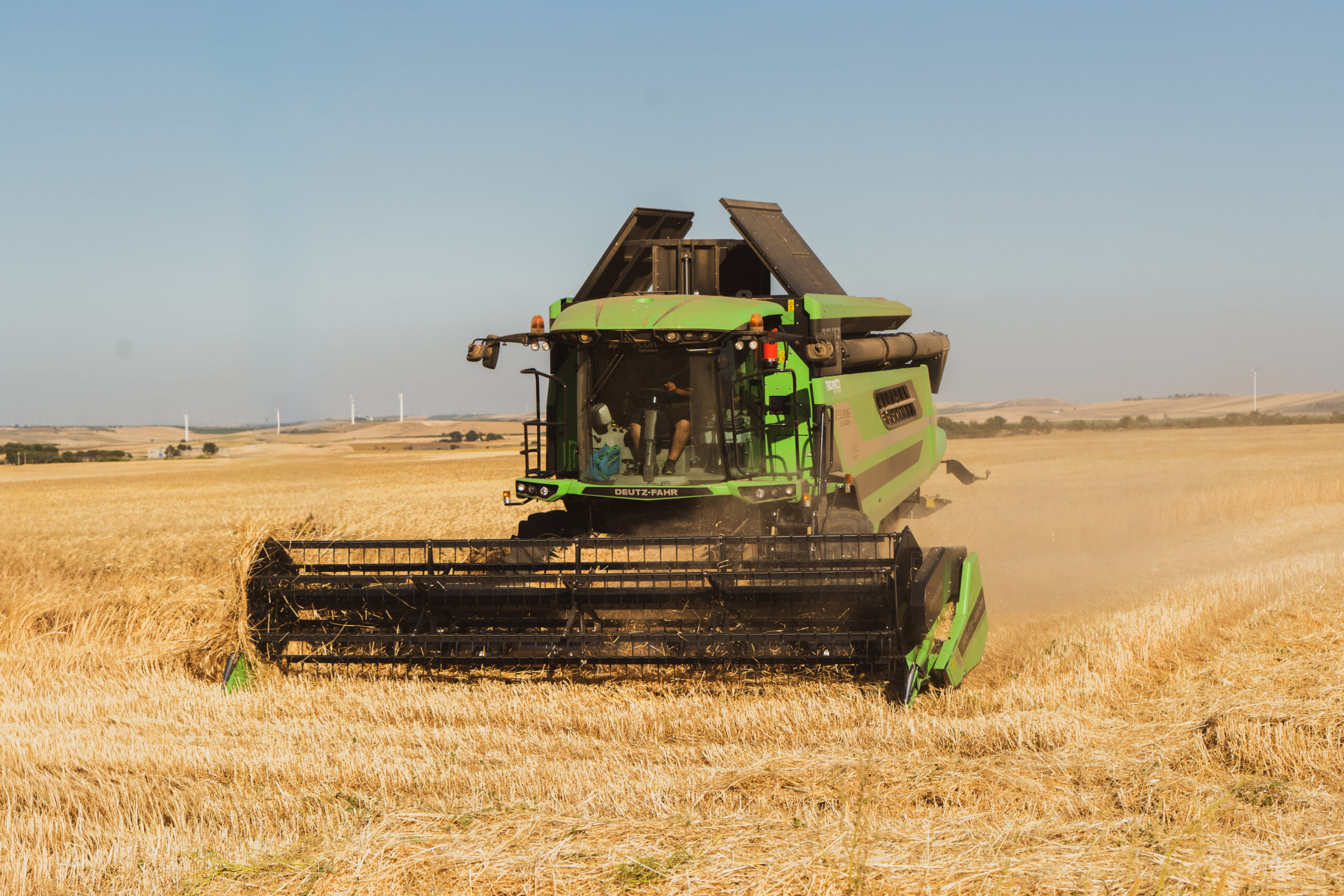 Green combine harvester at work in a golden grain field under a clear blue sky, dust rising behind it.