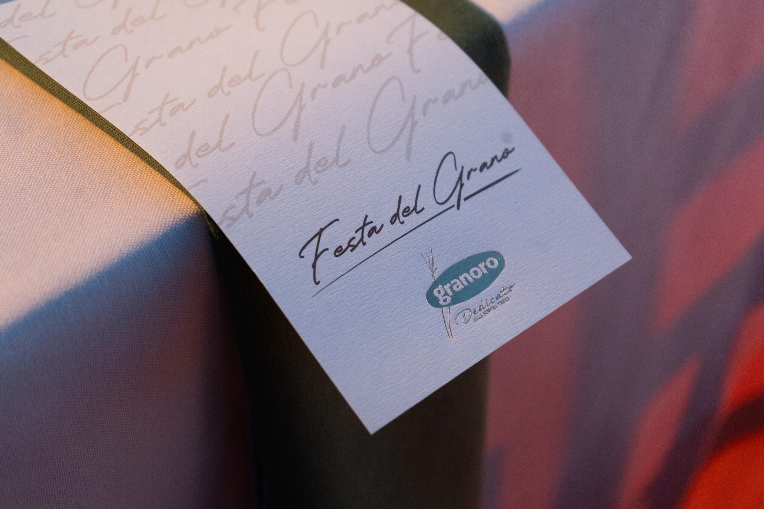 Close-up of a white card with cursive 'Festa del Grano' and a Granoro logo on textured fabric with a ribbon nearby.