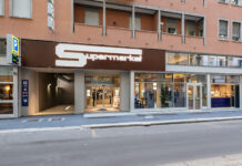 Brown storefront with large white 'S' logo and 'Supermarkt' sign; glass entrance with people inside, blue parking sign on left outside.