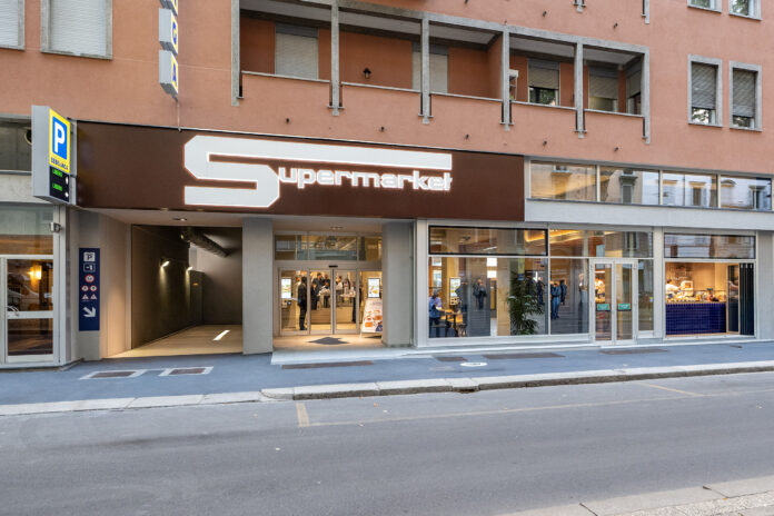 Brown storefront with large white 'S' logo and 'Supermarkt' sign; glass entrance with people inside, blue parking sign on left outside.