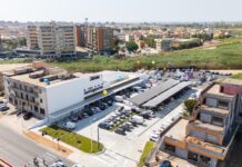 Tuttigiorni cresce nel Cagliaritano con il nuovo store di Quartu Sant’Elena Aerial view of a modern commercial building with a large parking lot and colorful balloons outside, surrounded by apartment blocks and green fields.