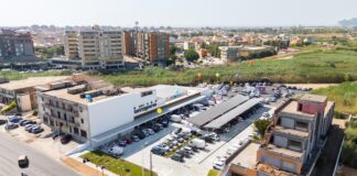 Aerial view of a modern commercial building with a large parking lot and colorful balloons outside, surrounded by apartment blocks and green fields.