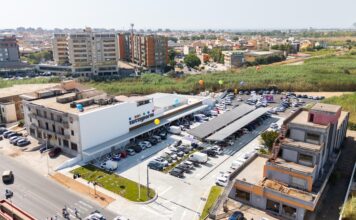 Tuttigiorni cresce nel Cagliaritano con il nuovo store di Quartu Sant’Elena Aerial view of a modern commercial building with a large parking lot and colorful balloons outside, surrounded by apartment blocks and green fields.