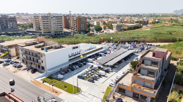 Foto Apertura Quartu CS (3) Aerial view of a modern commercial building with a large parking lot and colorful balloons outside, surrounded by apartment blocks and green fields.