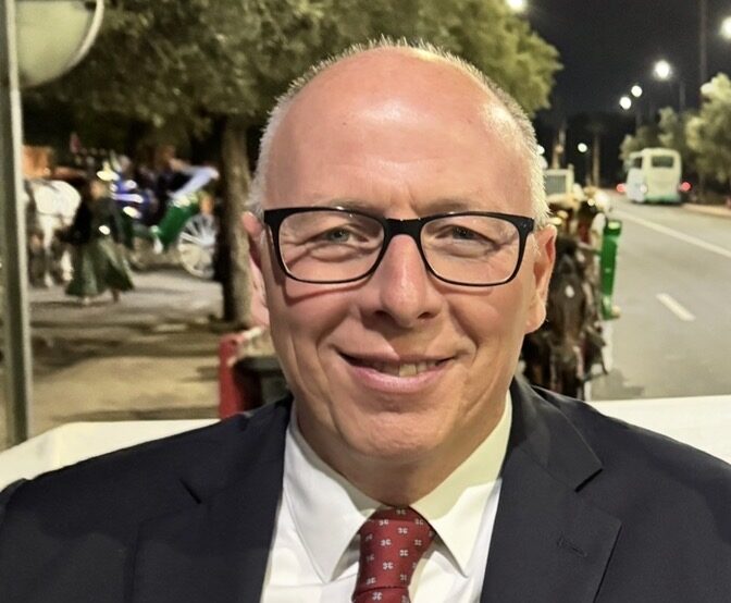 Smiling man in a dark suit and red patterned tie, wearing glasses, at night on a street.