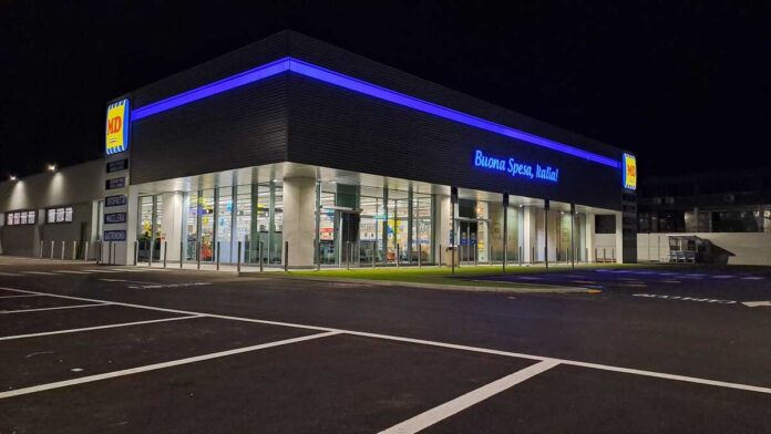 Foto_MD_Capaccio_2 Nighttime view of a modern MD supermarket with blue neon lighting and glass entrance, Italian slogan reading 'Buona Spesa, Italia!'