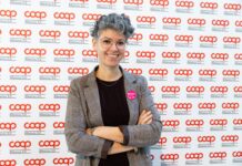 Smiling woman with short gray hair and glasses, arms crossed, wearing a plaid blazer at a Coop backdrop event with a pink badge on her lapel.