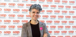 Smiling woman with short gray hair and glasses, arms crossed, wearing a plaid blazer at a Coop backdrop event with a pink badge on her lapel.