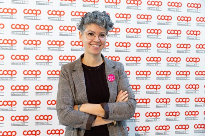 Francesca_Durighel Smiling woman with short gray hair and glasses, arms crossed, wearing a plaid blazer at a Coop backdrop event with a pink badge on her lapel.