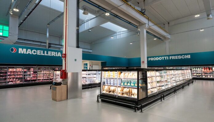 Interior of a modern supermarket with long refrigerated meat and dairy displays; teal signs read Macelleria and Prodotti Freschi on the walls.