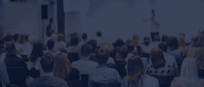 Audience seated in a conference hall watching a speaker presenting on stage at the front of the room.