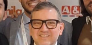 Dati, format e produttività: così Megamark prepara la nuova fase di sviluppo Smiling man in a dark suit and tie wearing black-framed glasses at a business event, group photo backdrop behind him.