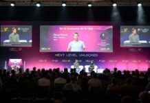 HIP sbarca in Italia: oltre 300 aziende e 400 relatori per l’ospitalità del futuro Two speakers on a stage during a conference, with large screens showing slides behind them and an audience in front,
