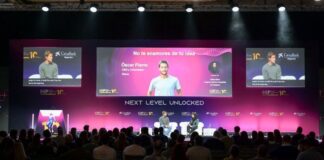 Two speakers on a stage during a conference, with large screens showing slides behind them and an audience in front,