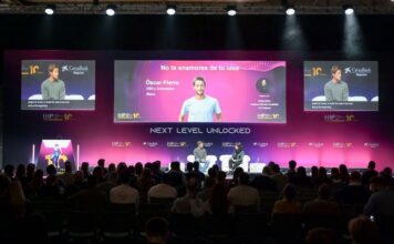 Two speakers on a stage during a conference, with large screens showing slides behind them and an audience in front,