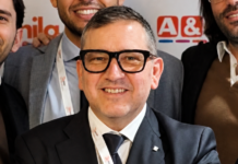 Dati, format e produttività: così Megamark prepara la nuova fase di sviluppo Smiling man in a dark suit and black-framed glasses posing for a group photo with colleagues in the background.