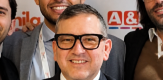 Smiling man in a dark suit and black-framed glasses posing for a group photo with colleagues in the background.