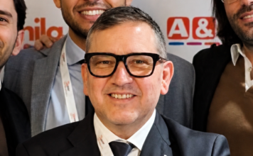 Dati, format e produttività: così Megamark prepara la nuova fase di sviluppo Smiling man in a dark suit and black-framed glasses posing for a group photo with colleagues in the background.