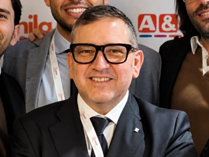 Maurizio-Strippoli-Gruppo Megamark-Responsabile-Controllo-di-Gestione Smiling man in a dark suit and black-framed glasses posing for a group photo with colleagues in the background.