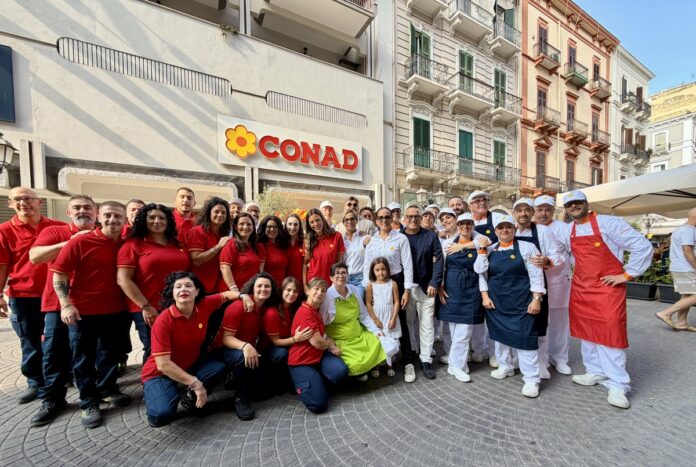 PDV_Taranto_1 Group of CONAD staff and chefs posing for a photo outside a supermarket on a sunny street.